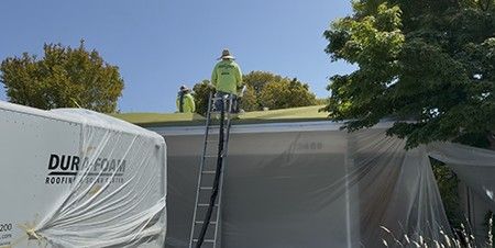 A worker on a ladder spraying polyurethane foam onto the roof, the wall below covered in plastic sheeting.