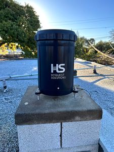 A dark blue HyQuest Solutions TB7 rain gauge on a concrete block on the roof, under a blue sky.