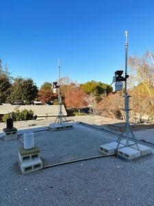 The gravel roof before the foam roof: two station masts on concrete blocks, each carrying an anemometer above its sensor suite, two rain gauges on blocks at left, autumn trees behind.