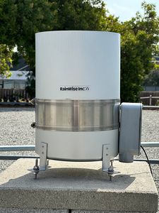 A RainWise industrial rain gauge: a tall gray cylinder on three leveling feet, on the roof beside a railing.