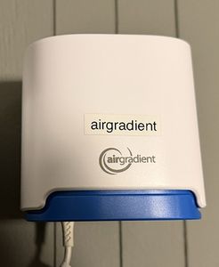 An AirGradient Open Air monitor: a cream wedge-shaped housing with a blue base, mounted on a wall.