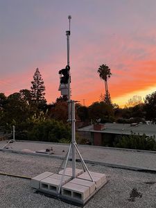 A Davis Vantage Pro2 Plus on a tripod mast on the gravel roof, photographed at sunrise against an orange sky.