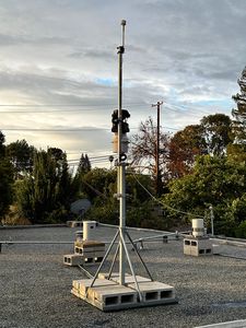 Two Davis stations on tripod masts on the gravel roof under a lightly clouded sky, rain gauges on blocks between them.