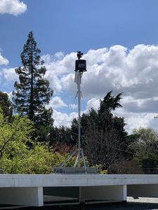 The RainWise Mark III-LR on a guyed mast above the roof parapet: wind vane and anemometer on top of a black rain collector, a white louvered radiation shield below, trees and a partly cloudy sky behind.